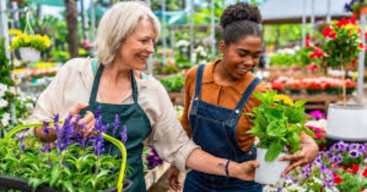  A person wearing gardening gloves holding a small, vibrant green seedling in a plastic nursery pot at a sunny outdoor garden center